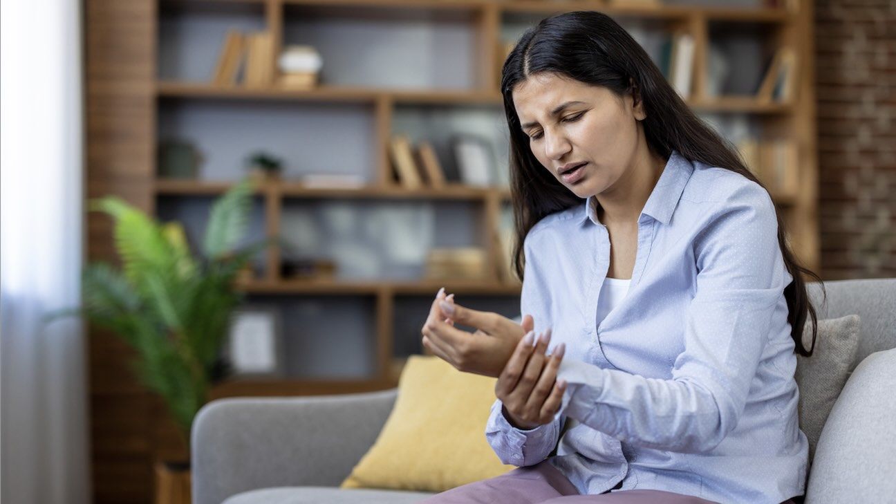 A woman with arthritis sits at home holding her wrist with a look of pain on her face