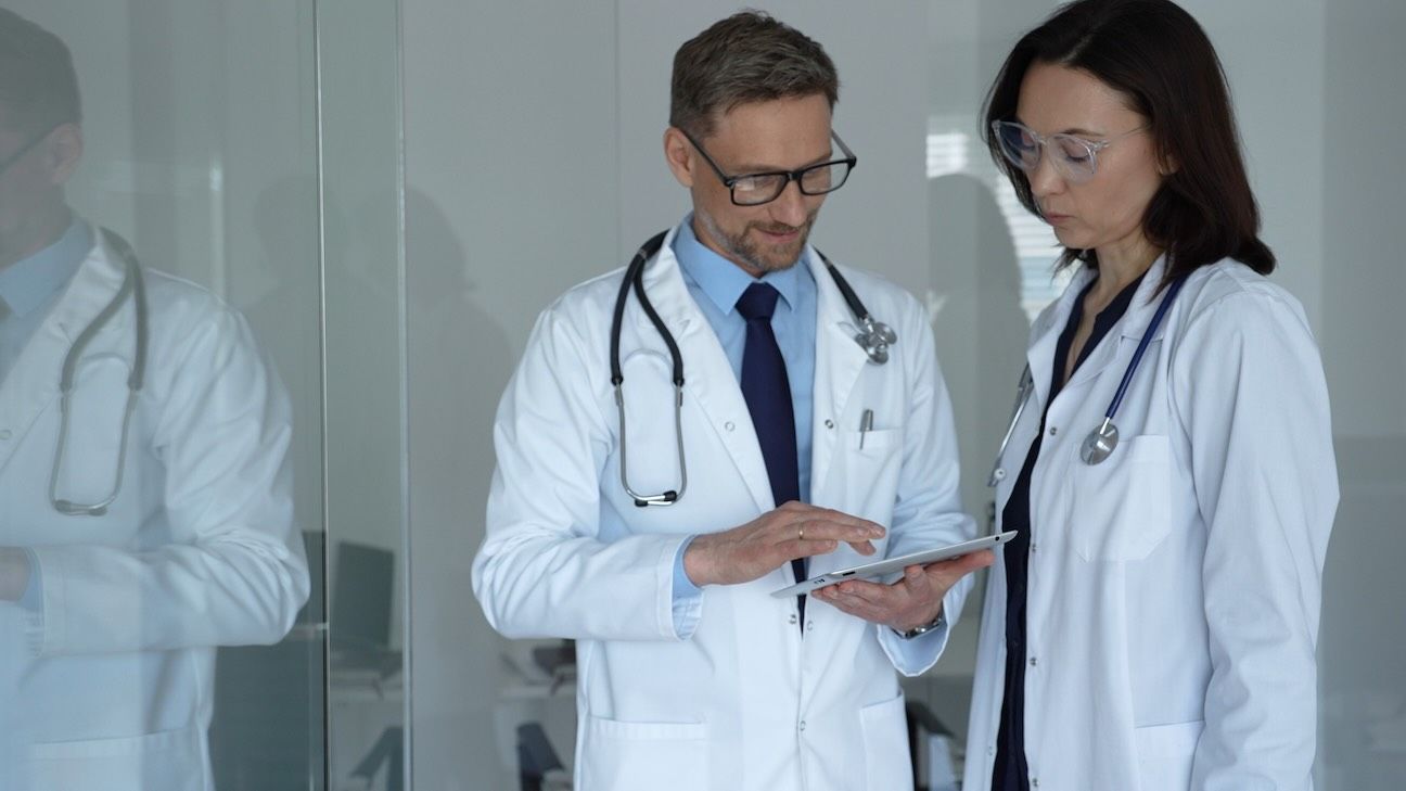 Male and female doctors collaborate over patient data on a tablet in a clinic, both in lab coats with stethoscopes, using digital tech for modern patient care and diagnosis