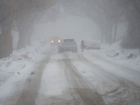 A pedestrian walks across 15th Avenue in Regina, battling the elements as a heavy snowfall hits the city. Vehicles drive slowly to manage reduced visibility.