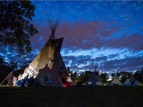 Teepees stand together at the Justice for our Stolen Children camp across from the Saskatchewan legislative building as summer daylight slowly gives way to a night sky over Regina. Vehicle headlights illuminate a "Justice for Colten" attached to the nearest teepee.