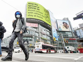 A pedestrian wearing a mask walks across Yonge Street at Dundas Square with a “Stop The Spread Of Covid 19” notice in the background during the Covid 19 pandemic, Friday April 3, 2020.