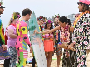 Tecumseh's Mark Micelli, left, in the bright body suit, holds a board as the Duchess of Sussex applies surfing wax during a visit by Prince Harry and his wife with members of a local surfing community group at Bondi Beach in Sydney on Oct. 19, 2018, during their Australian tour.