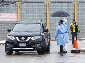 Health care workers use an umbrella to stay dry while speaking to people in their cars at the Carling Heights Optimist Community Centre COVID 19 assessment centre  in London, Ont. on Monday March 23, 2020. Fourteen cars were lined up a half-an-hour after it's 11:00am opening. (Derek Ruttan/The London Free Press)