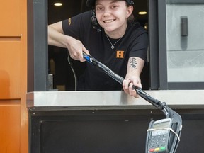 Jessica Kettle greets customers with a a smile and a payment machine at the end of a hockey stick while working the drive-through window at Harvey's on Highbury Avenue near Cheapside Street in London on Tuesday. The burger chain is using the iconic Canadian hockey stick to maintain a safe distance between employees and customers during the COVID-19 pandemic. (Derek Ruttan/The London Free Press)