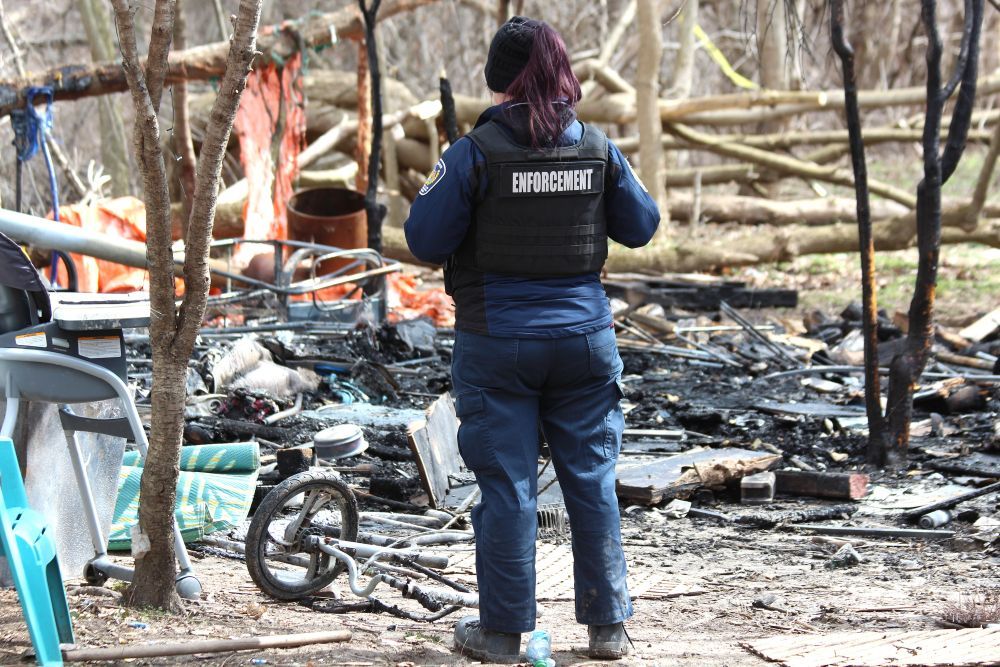 A bylaw officer surveys the damage from a fire at a homeless encampment in Watson Park in London. Photo taken on Monday April 7, 2025. (Dale Carruthers/The London Free Press)