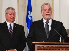 Quebec Premier Philippe Couillard gestures as he responds to media with Quebec Transport Minister Laurent Lessard, right, and Quebec Minister of Forests, Wildlife and Parks Luc Blanchette, left, after a cabinet shuffle at the legislature in Quebec City on Saturday, August 20, 2016.