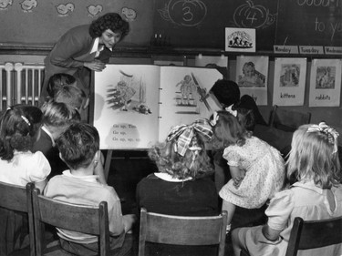 1946: Grade 1 students at Maisonneuve School, learn from an enormous book. Lots of pretty dresses and hair tied up with bows.