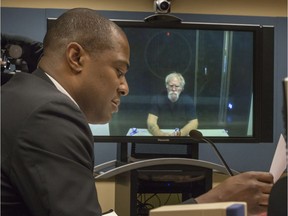 John Robert Boone, an American fugitive who was dubbed the Godfather of Grass because of run-ins with the law involving the large-scale production of marijuana, appears via closed circuit tv in the background from prison where he is being held, as CBSA's Anthony Lashley speaks at the Immigration and Refugee Board of Canada in Montreal on Thursday, Dec. 29, 2016.