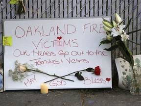 Signs and flowers adorn a fence near the site of a warehouse fire Monday, Dec. 5, 2016, in Oakland, Calif. The death toll in the Oakland warehouse fire on Friday climbed Monday with more bodies still feared buried in the blackened ruins, and families anxiously awaited word of their missing loved ones. Among the confirmed dead is singer Cash Askew of the band Them Are Us Too.