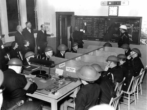 Photo dated March 27, 1943. Caption: If Montreal ever undergoes an air raid, the north division of the C.P.C. is ready to protect the district which spreads from Papineau to Hutchison and from the Laurier region to the Back River. It was shown at the official opening of the division headquarters in police station No. 21 last night. The imaginary raid conducted under the supervision of Chief Warden Victor Beaupre (... newsprint original is cut off...) white helmet at upper right, was attended by Director of Police Fernand Dufresne, Deputy Director C. Barnes and Frank V. Dowd, standing at rear. Mr. Dowd represented Montreal's public works director. The men seated at the table are C.P.C. workers who keep in constant touch with heads of local public services and utilities. On the blackboard is a record of service calls received by the division.