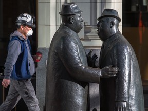 A worker wears a mask as he walks past the well-known statues of two businessman talking in downtown Calgary on Tuesday, March 17, 2020.