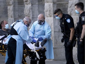 Montreal police officers watch as paramedics in full COVID-19 protective gear restrain a homeless man outside the temporary homeless shelter at the Bonsecours Market in Montreal, on Wednesday, May 20, 2020. The man was arrested after an altercation with a pedestrian passing the homeless shelter.