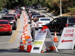 A woman wearing a face mask stands near people in cars lined up to be tested for COVID-19 as they make their way to a parking lot at Dodger Stadium amid the coronavirus pandemic on June 26, 2020 in Los Angeles, California.