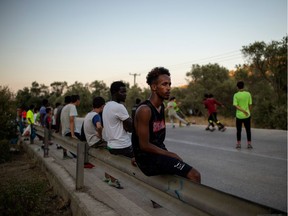 A migrant from Somalia sits on a road safety barrier as migrants play football outside of the makeshift camp next to the refugee camp of Moria, in the island of Lesbos on August 24, 2020. - Five years ago, the largest camp in Europe, slated to accommodate barely 2,770 people, was contained behind barbed wire. Asylum seekers disembarking on the northern coasts of the island, close to the Turkish shores, were just passing by, the time to register. And Moria was just a stopover on their way to Northern Europe. (Photo by ANGELOS TZORTZINIS / AFP)