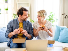 Happy couple sitting on sofa indoors at home, eating hamburgers.