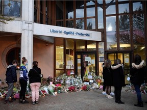 People stand in front of flowers at the entrance of a middle school in Conflans-Sainte-Honorine, 30 kms northwest of Paris, on October 17, 2020, after a teacher who had shown his students cartoons of the prophet Mohammed was decapitated by an attacker.