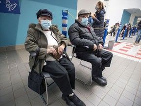 Annette Guimond and Maurice Bélisle take a seat while waiting in line outside the COVID-19 vaccination site at the Décarie Square on Monday.