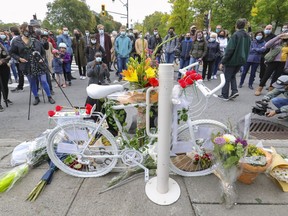 People gather around a bicycle painted white and decorated with flowers on the side of a street.