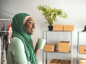 A Middle-eastern woman is indoors in her home office. She is wearing a head scarf. She is smiling behind her while sitting at her desk. She has coffee mugs in her hand.