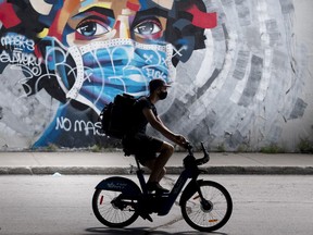 A man wears a face mask as he cycles by a mural defaced by graffiti objecting to mask usage, in Montreal, Sunday, Aug 30, 2020.
