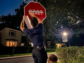 A man being hosted by another puts a sticker saying "Arrêt" on a "Stop" sign in a residential neighbourhood at night.