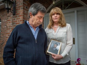 A man and woman stand on a front porch while she holds a photo of a young man in a picture frame