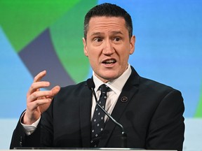 A white man wearing a suit is gesturing in front of a blue and green background.