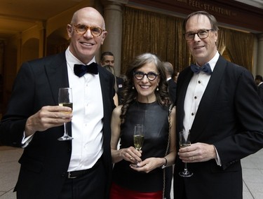 A woman in a black dress is flanked by two men in tuxedos at a party