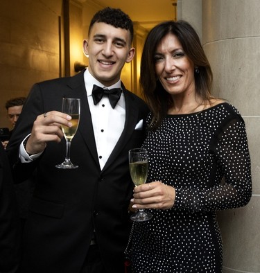 A woman in a black dress and a man in a tuxedo hold up wine glasses while being photographed at a ball