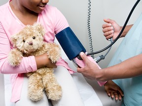 A Black child in a pink shirt holds a teddy bear while a doctor in blue scrubs checks her blood pressure in a doctor's office.