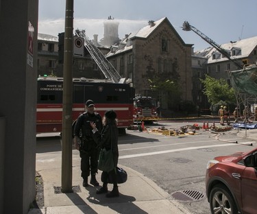 A woman talks to a police officer on the sidewalk as firefighters combat a blaze in the background
