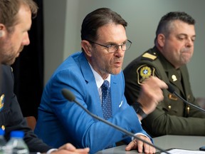 A politician in a blue suit gesticulates as he speaks while seated between two police officers at a press conference