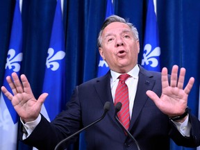 francois legault in a dark suit with red tie, hands extended, with quebec flags behind him