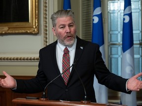 A man gesticulating as he speaks in front of two Quebec flags