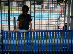 A young girl sitting on a bench looks at a pool through a fence