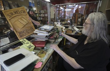 A young man shows the older man behind the cash in a store an old wooden sign that says "S.W. Welch Bookseller"