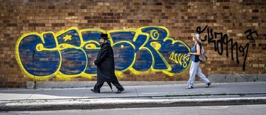 A Hassidic man in black garb and a woman in pants, shirt and sandals walk past yellow-blue graffiti tags on St-Viateur St.
