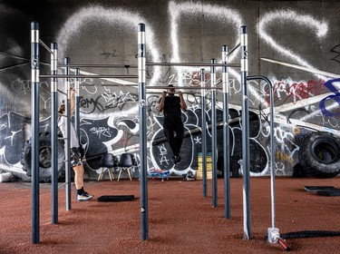 Two men do chinups on gym equipment, with graffiti on the back wall underneath the Bonaventure Overpass.