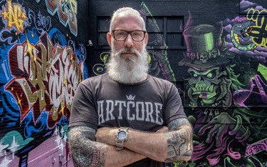 Grey-bearded Montreal city councillor Sterling Downey, with tattooed arms folded, standing in front of walls of bright graffiti.