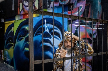 Visual artist Monk.E leans on a metal fence, with a mural he collaborated on with Cuban visual artist Mr. Myl in the background.