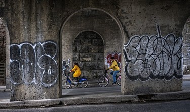 Two cyclists in yellow jackets, on Bixis, ride past graffiti on an Atwater Ave. underpass.