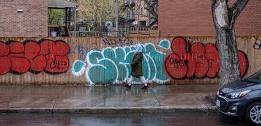 Pedestrian in a quilted coat walks past red and green graffiti on a brown fence along Atwater Ave. above Notre-Dame St.