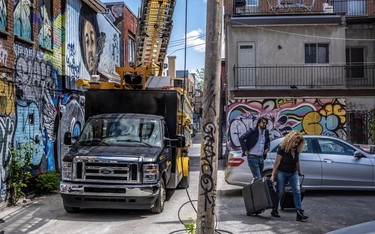 A man, with a woman dragging suitcases, walking in an alleyway along St-Laurent Blvd. in Montreal, framed by graffiti on building walls and a truck.