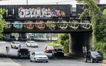 Cars drive under a railway overpass covered in tags as a railcar passes, also covered in tags, one marked 'JELOUS.'