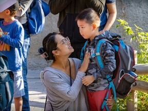 a woman kneels to talk to a young child with her hand on his chest. he has a large backpack and is looking toward fellow students