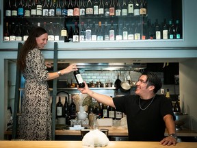 Lindsay Brennan is handing a bottle of wine to her husband, Juan Lopez Luna, at their restaurant, Alma. They are both behind a bar, with Lindsay standing on a ladder in front of a row of wine bottles on a shelf. Juan, who is to her right, has his arm outstretched to take the bottle.
