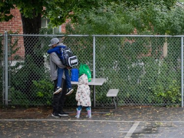 A young boy is held in the air by his father near a school yard fence while the boy's sister stands nearby in a rain jacket