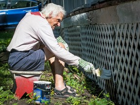 an elderly man sits on a stool to paint a fence