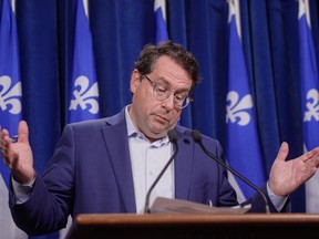 Quebec Education Minister Bernard Drainville gestures with both hands up as he speaks in front of a bunch of Quebec flags.