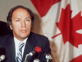 A man wearing a suit with a rose in his button hole speaks into a number of microphones in a scene from the 1970s. There's a Canadian flag behind him.